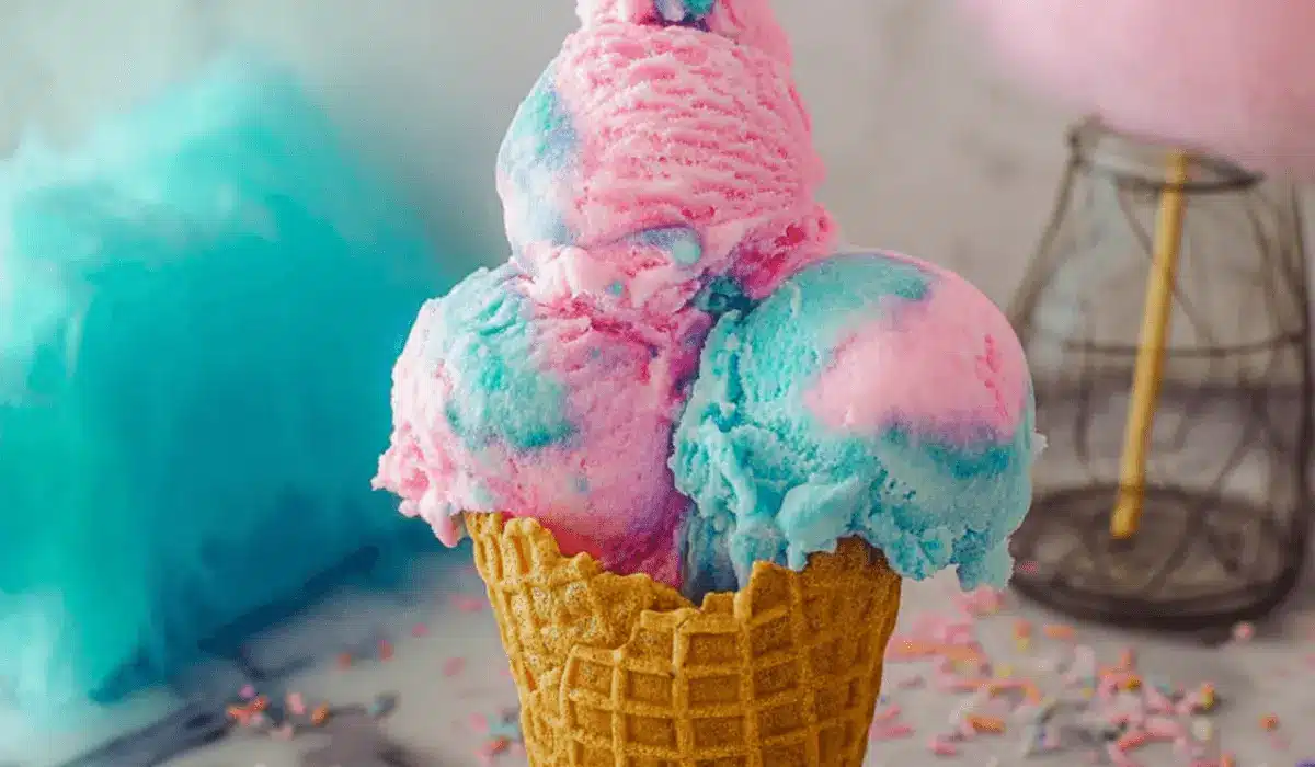 Close-up of cotton candy ice cream in a waffle cone with pink and blue scoops against a carnival-style background