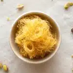 Flatlay of crispy kataifi nests in a bowl with pistachios and chocolate pieces on a white cloth