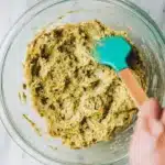 An overhead shot of hands using a teal spatula to gently fold toasted kataifi into the creamy pistachio base in a glass bowl.